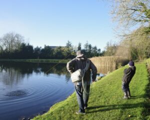 A young angler plays a fish on his fly rod while a man approaches him to net it. 