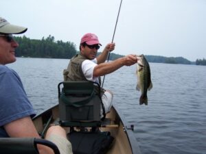 A fisherman holds up a bass and his fishing rod while sitting on a boat in the middle of the lake