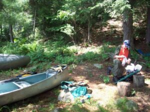 A man sits beside his canoes and outdoor equipment. 