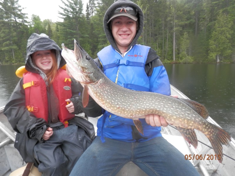 Two young, happy anglers sit beside one another on a boat holding out a northern pike.