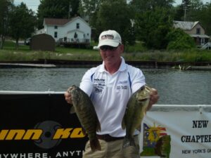 A competition angler holding up two bass while out fishing on a boat.