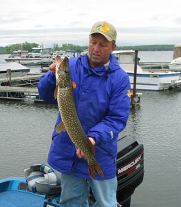 A middle-aged man holds out a pike he caught while fishing from a boat dock.