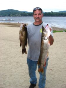 A fisherman holds up a bass in his right hand and a northern pike in his left hand. 