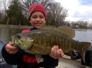 A child holds a massive smallmouth bass, posing for the camera with a big smile.