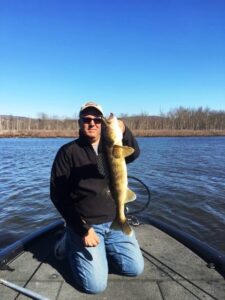 A walleye held upright by a fisherman at the hull of his boat surrounded by a lake and woodland.