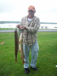 An older man holding two pike upright on a stringer with a lake in the background.