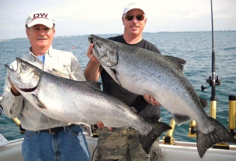 Two men stand on a boat, both holding large salmon. 