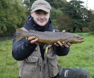 A fisherman holds out a brown trout with some green scenery in the background. 