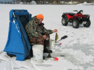 A happy fisherman holds up a yellow perch while out ice fishing on a lake with his quad and ice shelter.