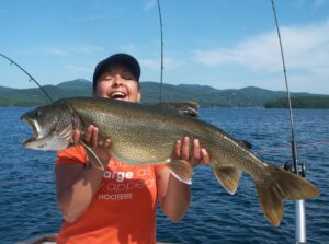 A joyful angler holds up a huge lake trout with three rods in the background and a beautiful lake view. 