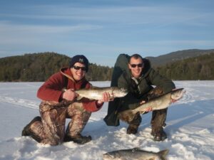Two anglers hold out lake trout during ice fishing while one lies in the snow beneath them.