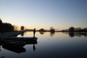 A fisherman stands on the dock at a lake as the sun sets. 