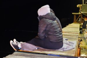 Two fishermen sit beside their fishing rods in the dark dressed in comfortable clothing. 