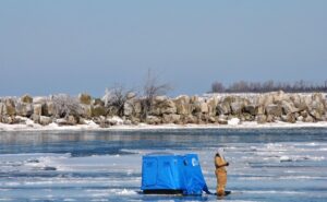 A fisherman stands ice jigging on a frozen lake beside his snow shelter.