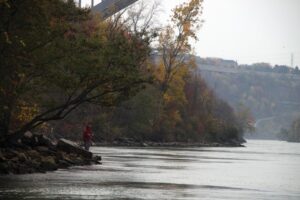 A fisherman stands on the shore of a river.